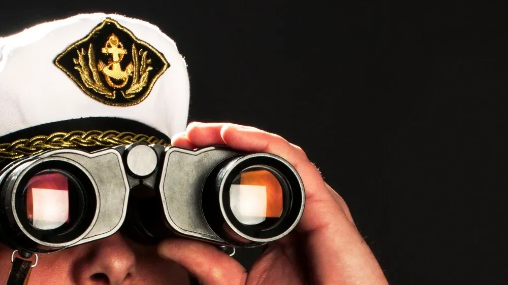 A crew member in a captain's hat looking through binoculars